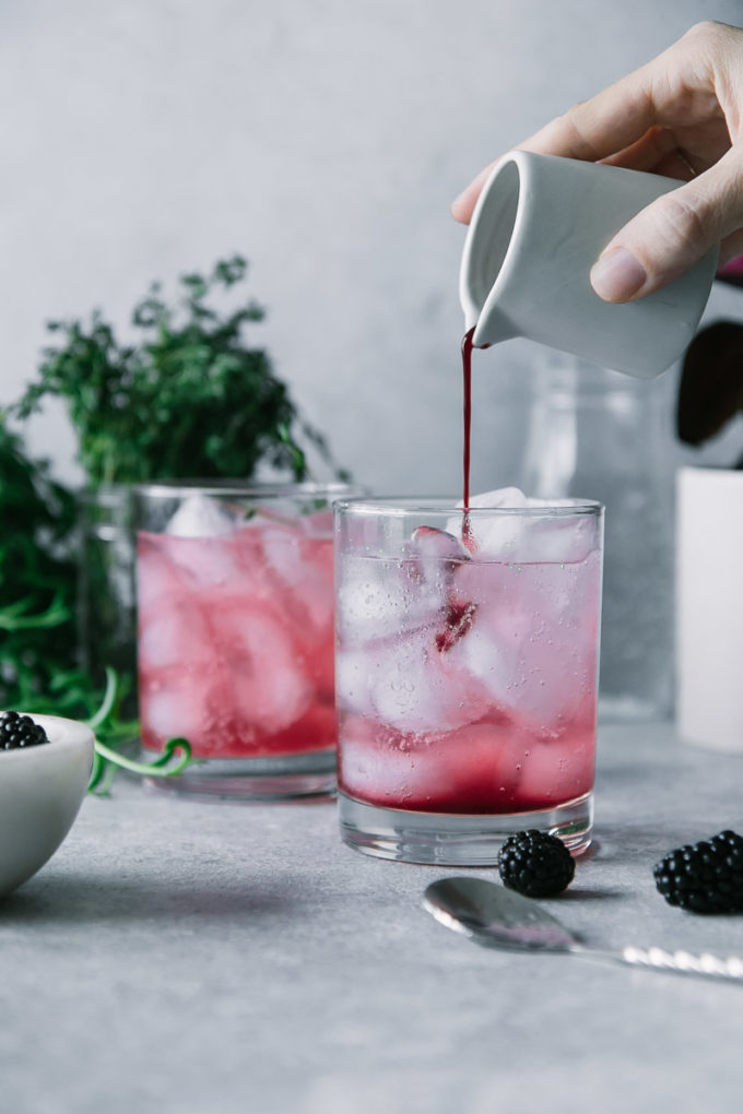 a hand pouring blackberry juice into a glass of sparkling water with the words "Blackberry Thyme Spritzer" in black writing