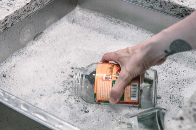 a hand submerging an empty glass food jar with a label into a sink filled with soapy water