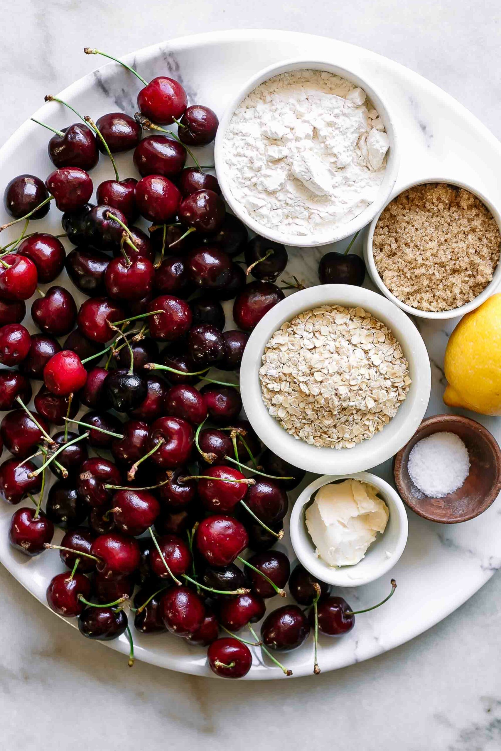 fresh cherries and bowls of flour, oats, sugar, butter, salt, and a lemon on a white table