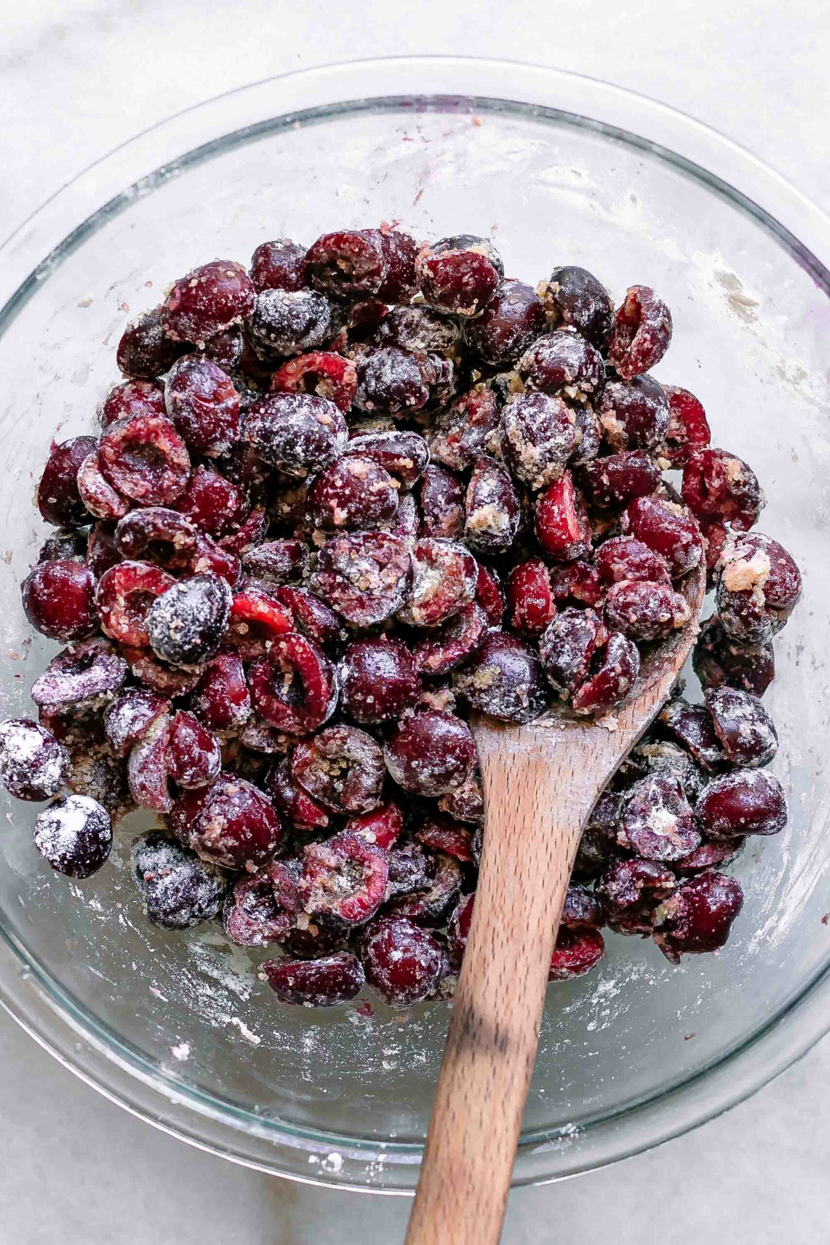 cut cherries, sugar, and flour mixed in a glass mixing bowl with a wooden spoon