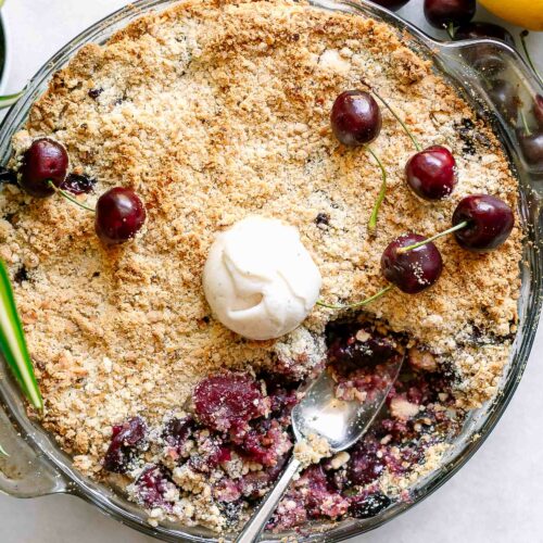a baking dish with a vegan cherry crisp topped with fresh cherries and a scoop of ice cream on a white table