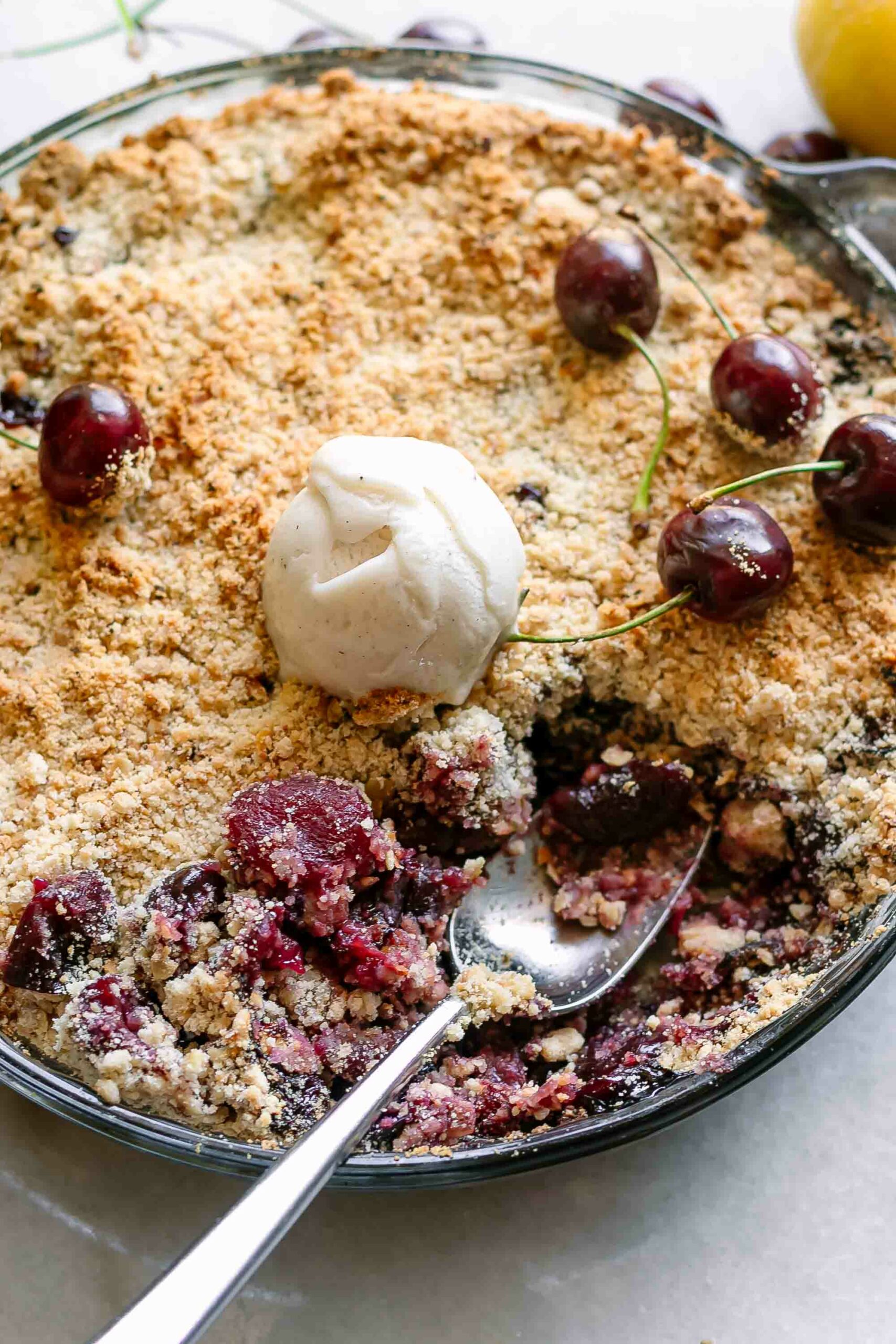 a close up of a gold spoon inside a baked cherry crumble in a baking dish on a white table